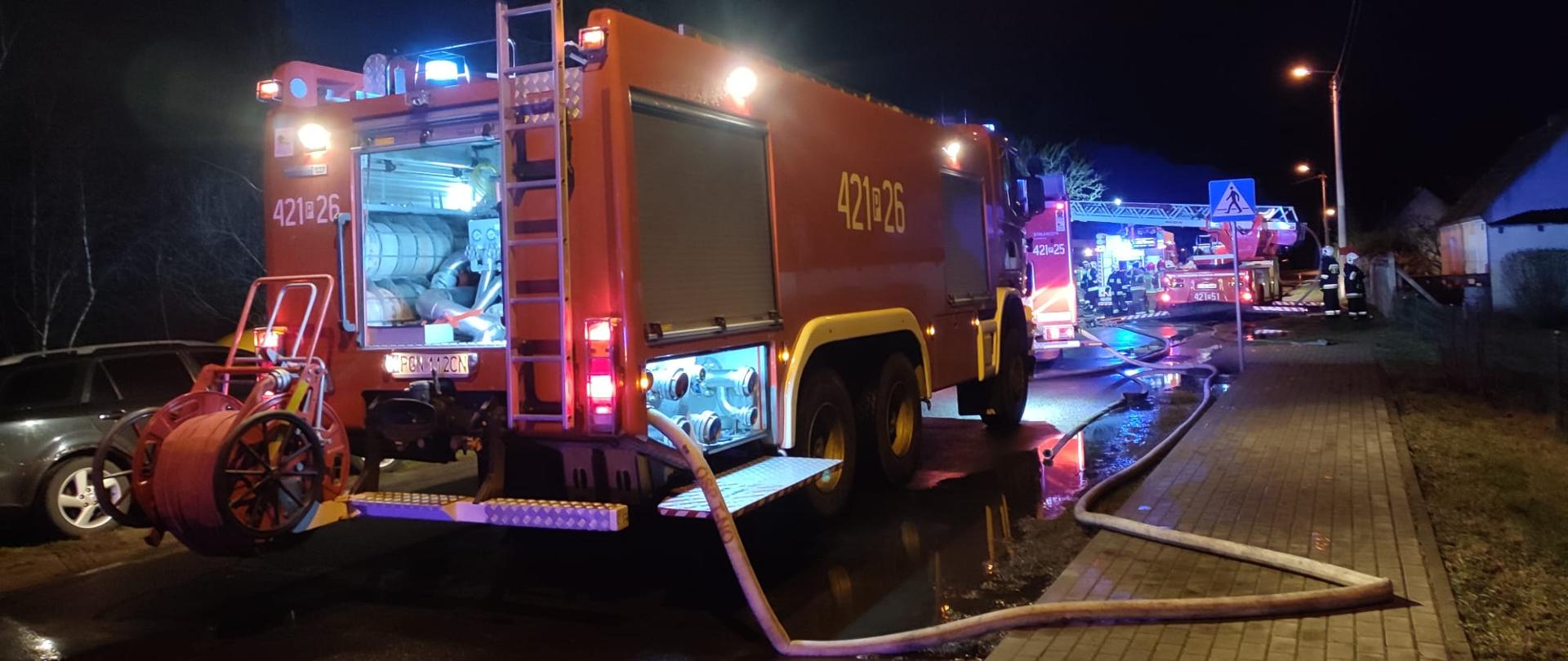 Na zdjęciu widać samochody pożarnicze, które stoją na drodze asfaltowej. Od samochodów rozwinięte są linie gaśnicze. W tle widać drabinę mechaniczną przygotowywaną do działań