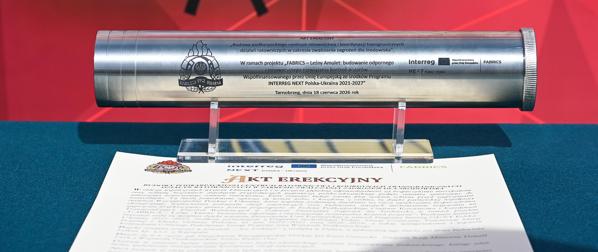 Metalowa, srebrna tuba na akt erekcyjny spoczywa na przezroczystej podstawce, umieszczonej na stole przykrytym ciemnozielonym obrusem. Przed tubą leży rozłożony dokument z nagłówkiem „AKT EREKCYJNY”. W tle widoczna jest duża czerwona ścianka z oficjalnym, niebiesko-złotym godłem i napisem „PAŃSTWOWA STRAŻ POŻARNA”.