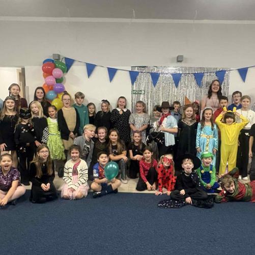 Duża grupa dzieci w różnym wieku pozuje w sali udekorowanej chorągiewkami, srebrnym tłem i balonami. Wiele osób ma na sobie przebrania — od bajkowych sukienek po stroje zwierząt, bohaterów czy postaci z gier. Dzieci stoją i siedzą w kilku rzędach, uśmiechają się. Biorą udział szkolnej zabawie andrzejkowej.