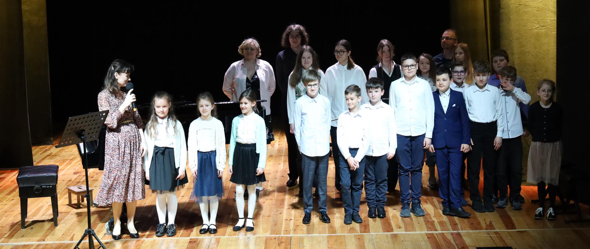Fotografia wspólna wszystkich wykonawców koncertów dla uczniów szkół podstawowych. Ustawieni w rzędzie na scenie MOK w Głubczycach, przed oraz z prawej strony obok fortepianu.