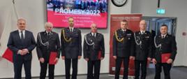 Zdjęcie zrobione w siedzibie Komendy Wojewódzkiej PSP w Gdańsku. W rzędzie stoi 6 strażaków w galowym mundurze oraz pan w garniturze. Trzech strażaków z OSP trzyma czerwone teczki. Stoją oni na tle flagi Polski oraz telebimie z napisem "PROMESY 2022 Komenda Wojewódzka Państwowej Straży Pożarnej w Gdańsku"