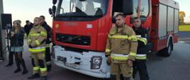 samochód pożarniczy obok niego czterech strażaków ochotników ubranych w umundurowanie bojowe po lewej stronie goście