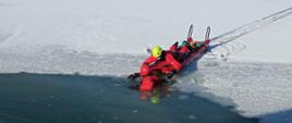 Zdjęcie przedstawia dwóch strażaków. Jeden na lodzie porusza się saniami lodowymi i podejmuje z wody drugiego strażaka. Obaj ubrani i wyposażeni są w ubrania i sprzęt przeznaczony do ratownictwa wodno-lodowego.
