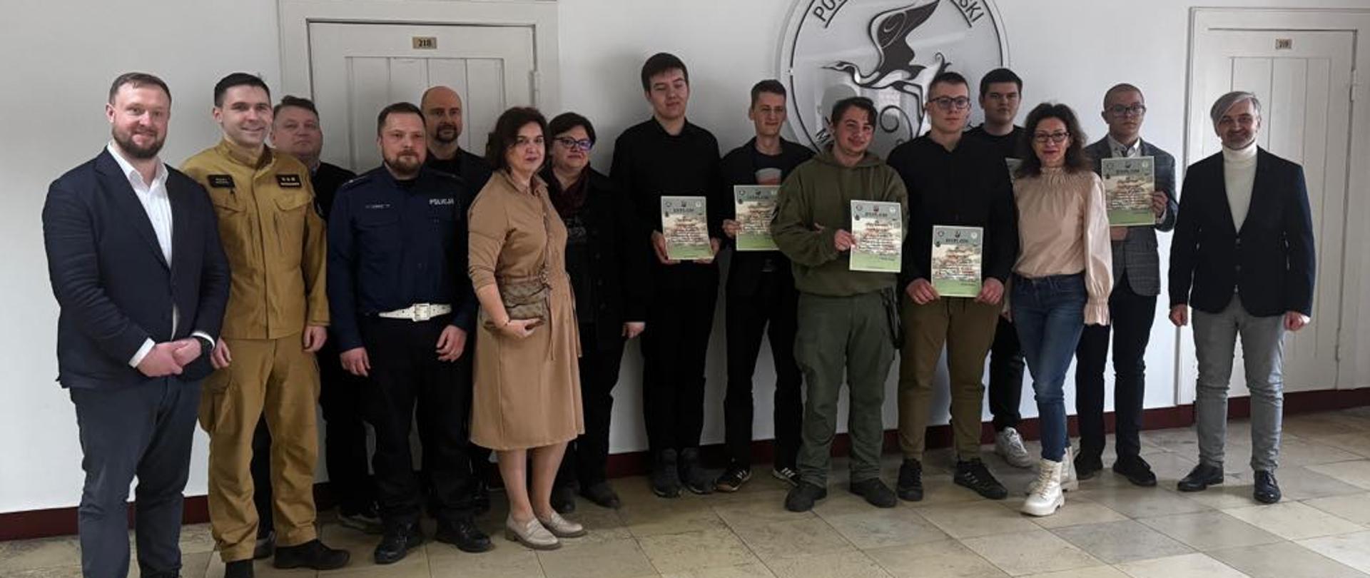 na zdjęciu na korytarzu stoi w rzędzie piętnaście osób na tle białej ściany, podłoga z beżowej terakoty