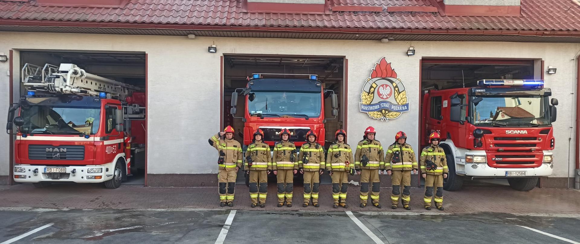 Na zdjęciu widać plac komendy PSP w Ustrzykach Dolnych. Przed garażami w których bramy są podniesione widać trzy samochody ciężarowe Państwowej Straży Pożarnej. Przed pojazdami stoi ośmiu funkcjonariuszy straży pożarnej w ubraniach specjalnych. 