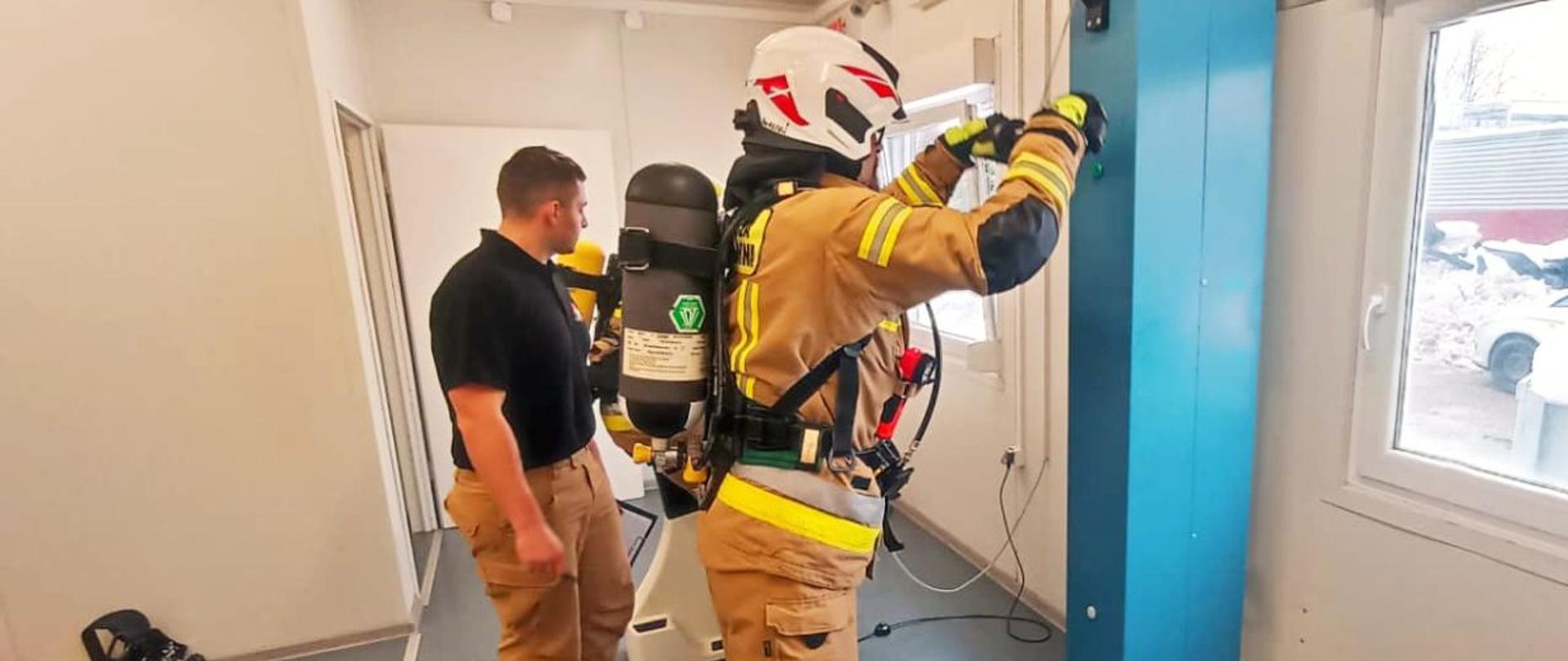 Zdjęcie przedstawia strażaka OSP wykonującego ćwiczenia "młot" przed wejściem do komory dymowej. Obok stoi funkcjonariusz PSP.