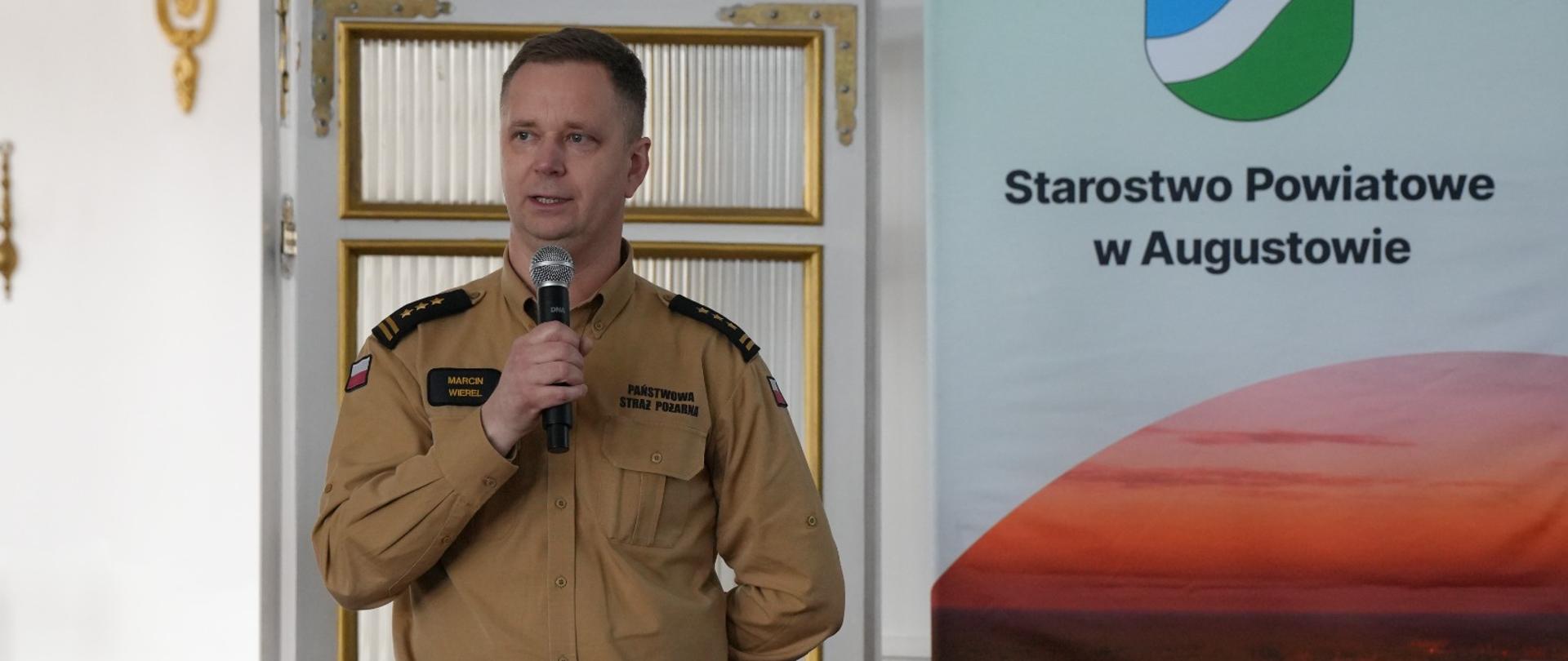 Augustowska konferencja dot. realizacji Programu Ochrony Ludności i Obrony Cywilnej - st. bryg. Marcin Wierel podczas swojego wystąpienia , trzyma mikrofon, stoi, a za nim baner starostwa powiatowego w Augustowie