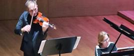 Mężczyzna stojący przy pulpicie gra na skrzypach, obok po prawej stronie kobieta gra na fortepianie - zbliżenie.
