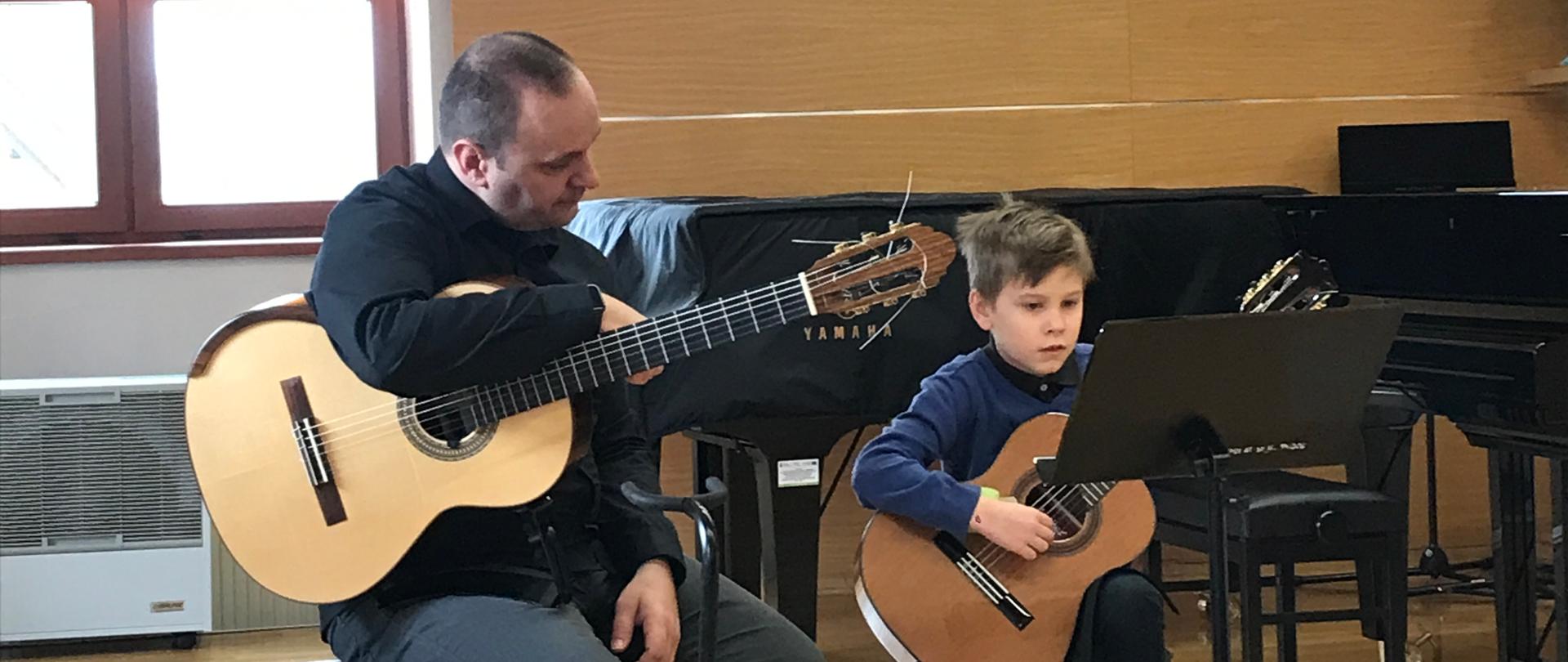 Na scenie sali kameralnej prowadzący warsztaty trzymający gitarę na kolanach siedząc, obok niego uczeń również siedzi, nogę ma na podnóżku i gra wpatrzony w nuty, które są ułożone na pulpicie ustawionym przed nim. Za nimi z lewej strony okno, a pod nim nawilżacz powietrza. Z tyłu za nimi i po prawej stronie dwa fortepiany oraz ława fortepianowa
