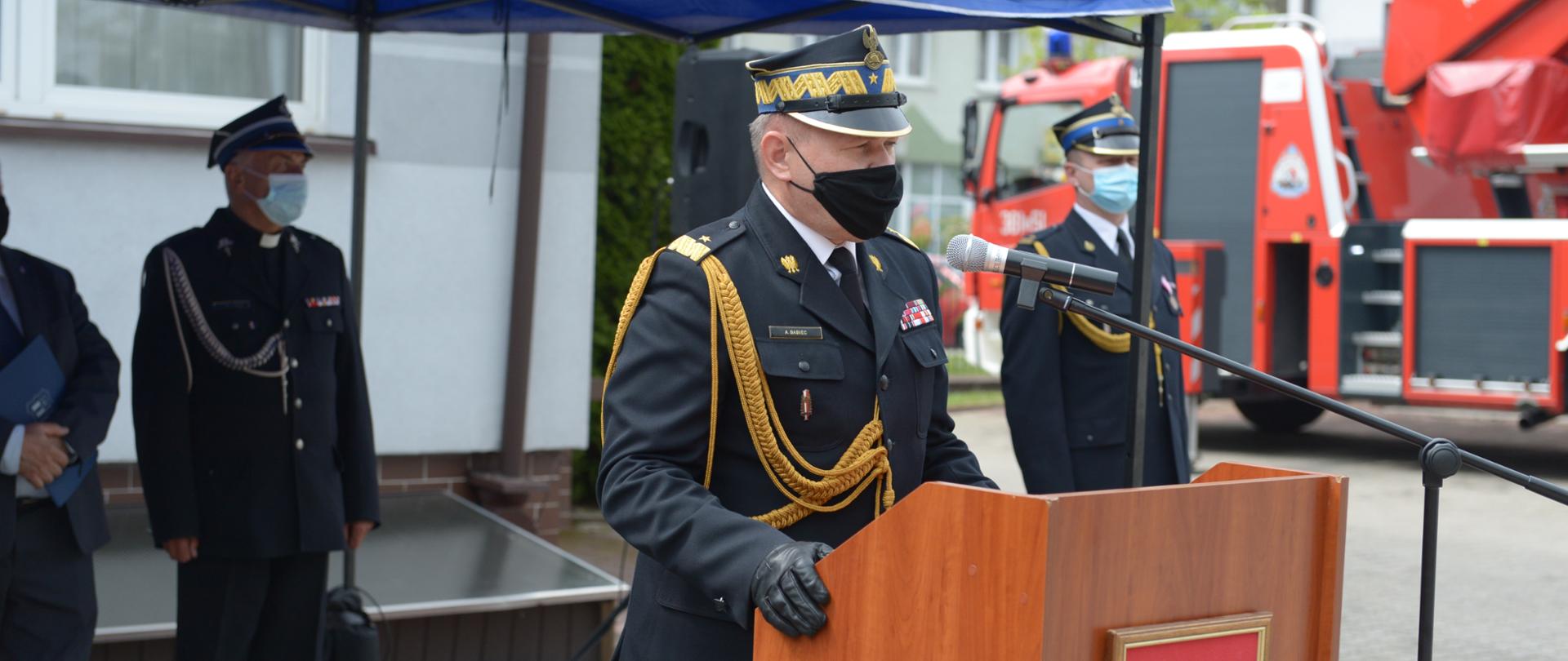Przed mównicą z mikrofonem stoi strażak - generał i przemawia. Za mównicą ustawiony pawilon, pod którym stoi strażak w umundurowaniu oraz mężczyzna w garniturze. 