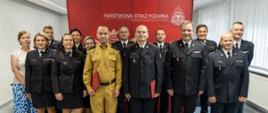Na tle czerwonego baneru z godłem Polski i napisem „PAŃSTWOWA STRAŻ POŻARNA KOMENDA WOJEWÓDZKA PSP W OPOLU” stoi grupa funkcjonariuszy w mundurach. W centralnej części zdjęcia wyróżniają się osoby: ubrana w żółty, piaskowy mundur służbowy, trzymająca czerwoną teczkę – rotę ślubowania oraz w umundurowaniu wyjściowym trzymająca czerwoną teczkę z pochwałą. Pozostali uczestnicy uroczystości, zarówno kobiety, jak i mężczyźni w galowych mundurach PSP. Cała scena oddaje powagę i rangę wydarzenia.