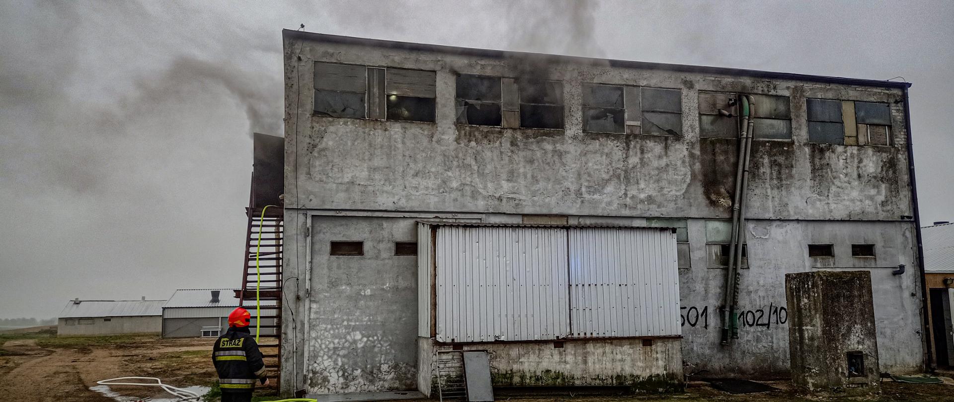 Na fotografii znajduje się piętrowy budynek gospodarczy. Z jego okien wydobywa się dym. Po jego lewej stronie znajdują się schody, po których poprowadzona jest linia gaśnicza. Przed schodami znajduje się strażak. W oddali znajdują się następne budynki gospodarcze.