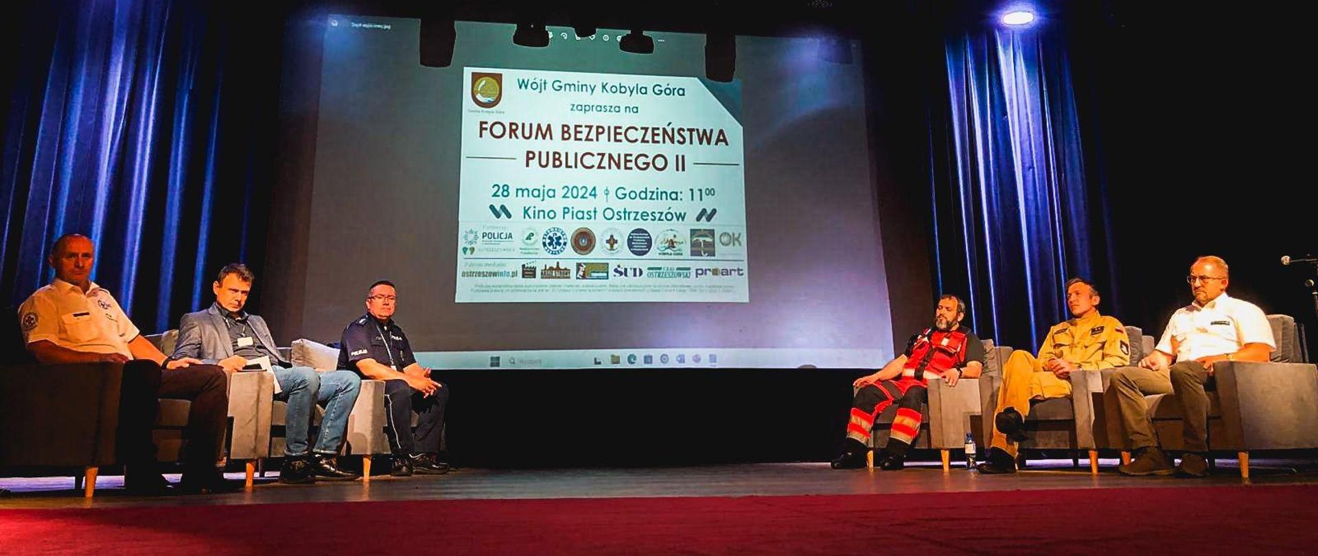 Prelegenci siedzą na fotelach na scenie. W tle prezentacja multimedialna