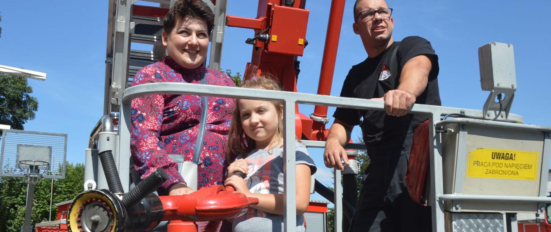 Dzień ze strażakami. Pani Magdalena i Alicja znajdują się w koszu podnośnika hydraulicznego. Towarzyszy im strażak. Alicja "operuje" działkiem. W tle drzewa. 