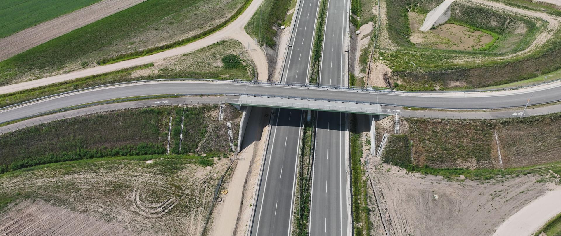 Widok z lotu ptaka na dwujezdniową drogę ekspresową przebiegającą pod wiaduktem, który przecina ją pod niewielkim kątem; wokół znajdują się pola uprawne, drogi dojazdowe oraz pasy zieleni.
