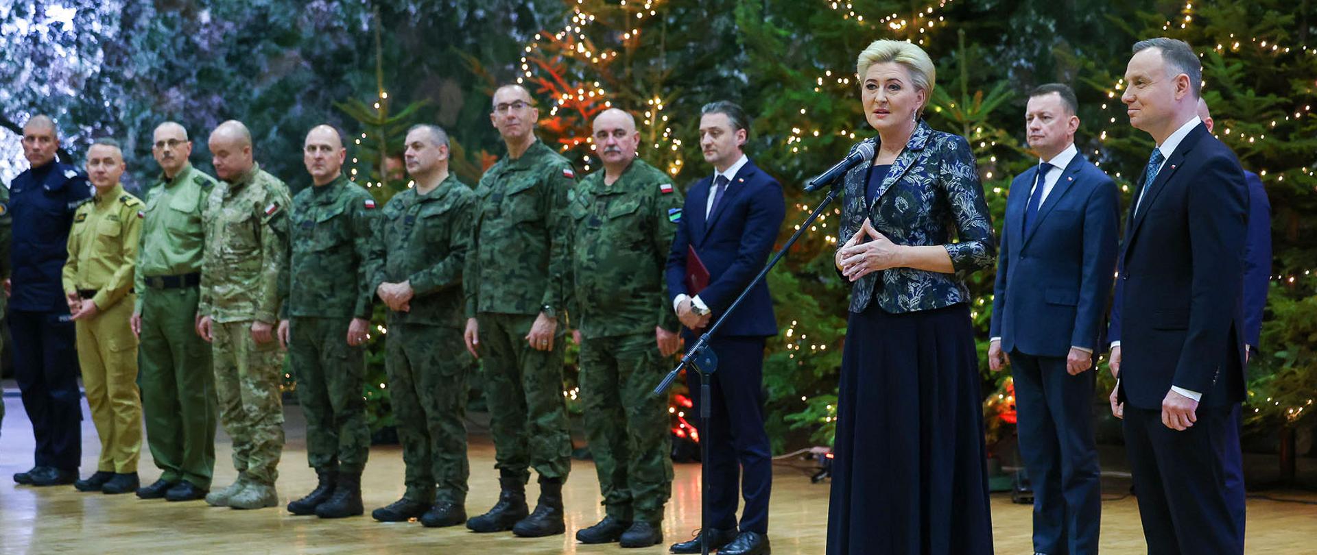 Do mikrofonu przemawia Pierwsza Dama, obok niej stoi Prezydent RP, w tle stoją ministrowie i szefowie służb mundurowych