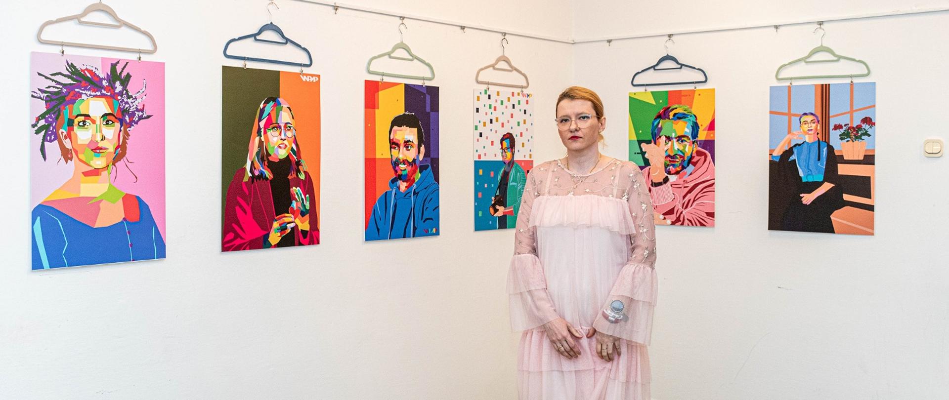 Zdjęcia przedstawia słuchaczkę stojącą na tle zdjęć, które są zawieszone na białej ścianie. Ubrana jest w jasną sukienkę. 