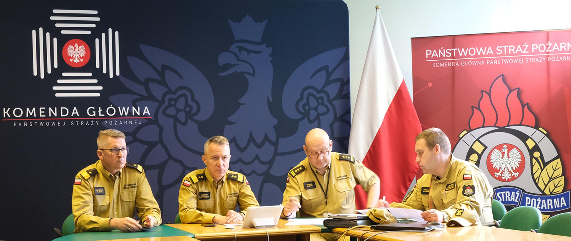 Czterej mężczyźni w mundurach Straży Pożarnej siedzących przy stoliku w trakcje wideokonferencji. W tle flaga państwowa oraz ścianki z logotypami Państwowej Straży Pożarnej oraz Komendy Głównej Państwowej Straży Pożarnej
