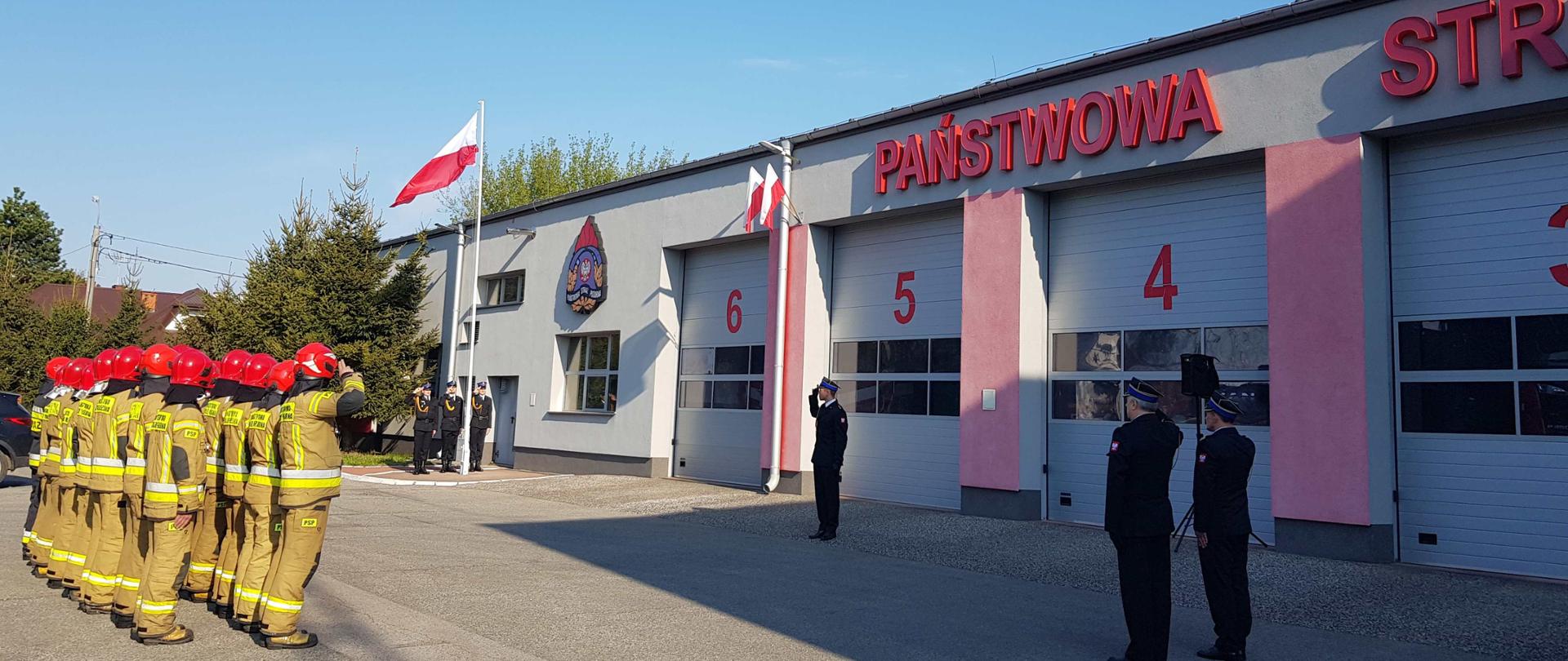 W tle poczet flagowy z trzech salutujących strażaków stoi przy maszcie na którym wciągnięta jest flaga RP, budynek strażnicy z widocznym logo PSP i napisem Państwowa Straż. Na pierwszym planie Komendant Powiatowy z Zastępcą i pododdział strażaków. 