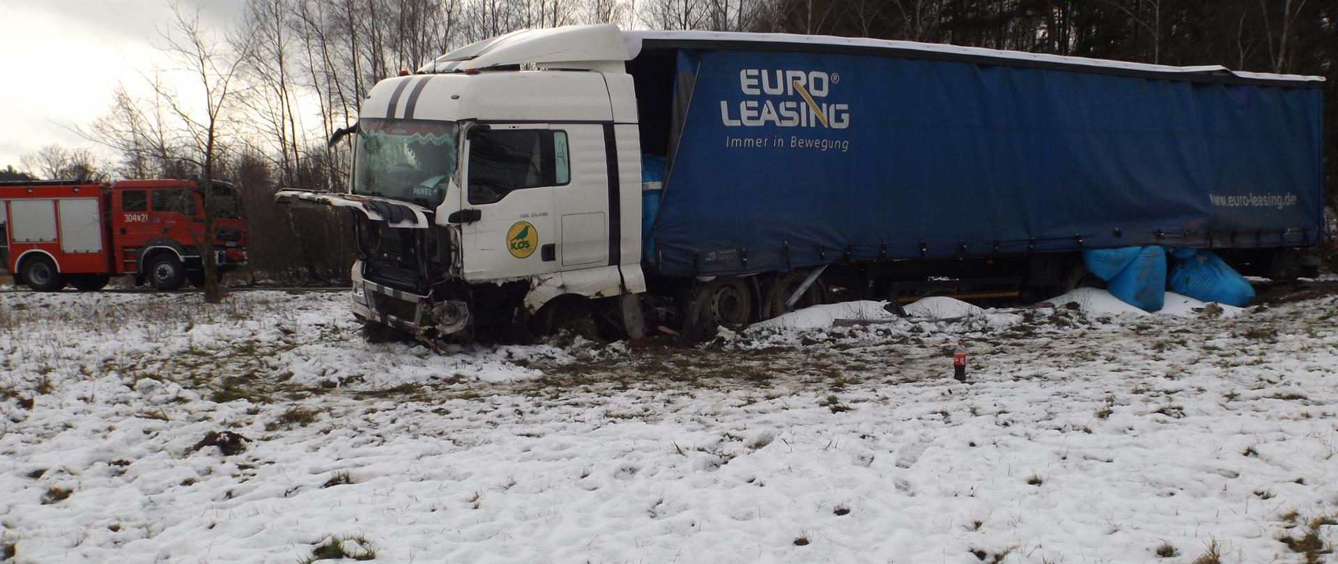 Zdjęcie przedstawia samochód ciężarowy biorący udział w zdarzeniu - ma uszkodzoną kabinę oraz granatową naczepę spod której wypadają niebieskie worki. Z lewej strony ciężarówki znajduje się samochód straży pożarnej.