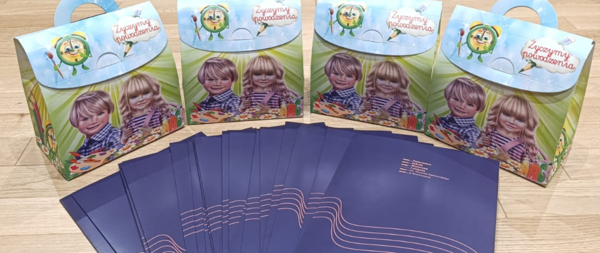 Na zdjęciu pudełka z rączką. Na pudełko uśmiechnięty chłopiec, a obok niego dziewczynka- blondynka. Przed dziećmi farby z pędzlami. Na niebieskim zamknięciu pudełka zielony zegar z pędzlem oraz napis: "Życzymy powodzenia". Przed pudełkami leżą granatowe teczki z logiem szkoły.