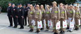 dwa pododdziały po lewej złożony z druhów po prawej z dzieci z młodzieżowej drużyny pożarniczej w tle drzewa