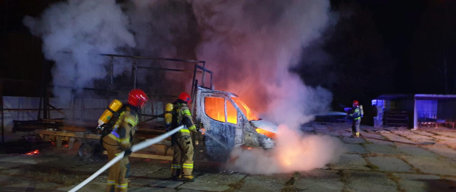 Na zdjęciu widzimy strażaków gaszących palący się samochód dostawczy Iveco. Strażacy ubrani w sprzęt ochrony układu oddechowego podają prąd wody na palący się samochód osobowy, z samochodu wydobywa się ogień i gęsty dym.