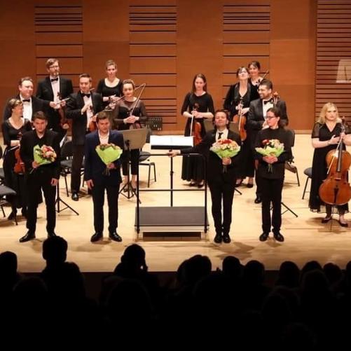 Zdjęcie przedstawia salę koncertową Filharmonii Kameralnej im. Witolda Lutosławskiego w Łomży. Na zdjęciu widzimy orkiestrę z instrumentami. Przed orkiestrą stoi trzech solistów oraz dyrygent. 