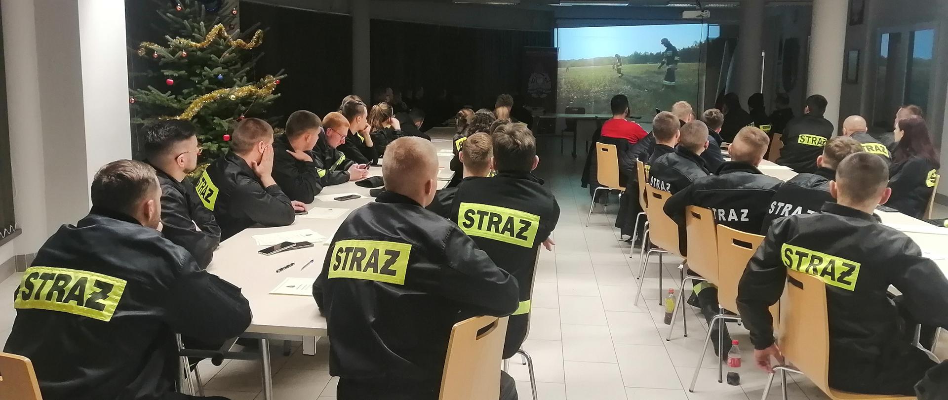 Kilkudziesięciu mężczyzn ubranych w czarne ubrania koszarowe strażackie z odblaskowymi paskami, czarnym butami, siedzą w ławkach i oglądają wyświetlany film. W tle wyświetlany jest film na ekranie..