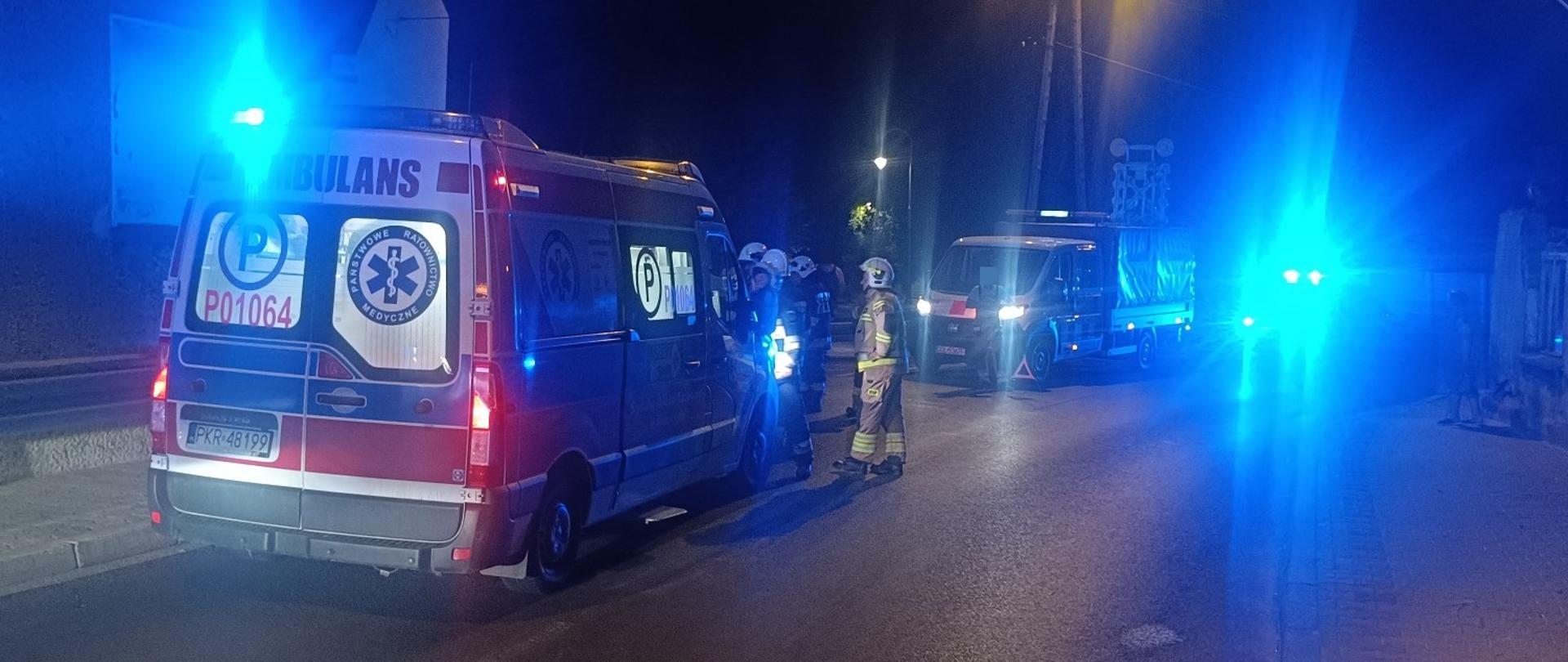 Zdjęcie przedstawia strażaków oraz karetkę pogotowia ratunkowego na miejscu wypadku w godzinach wieczornych.