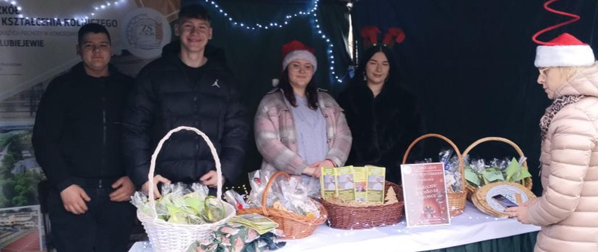 Widok na całe szkolne stoisko z koszami upominków, w głębi młodzież reprezentująca szkołę, obok pani widecyrektor