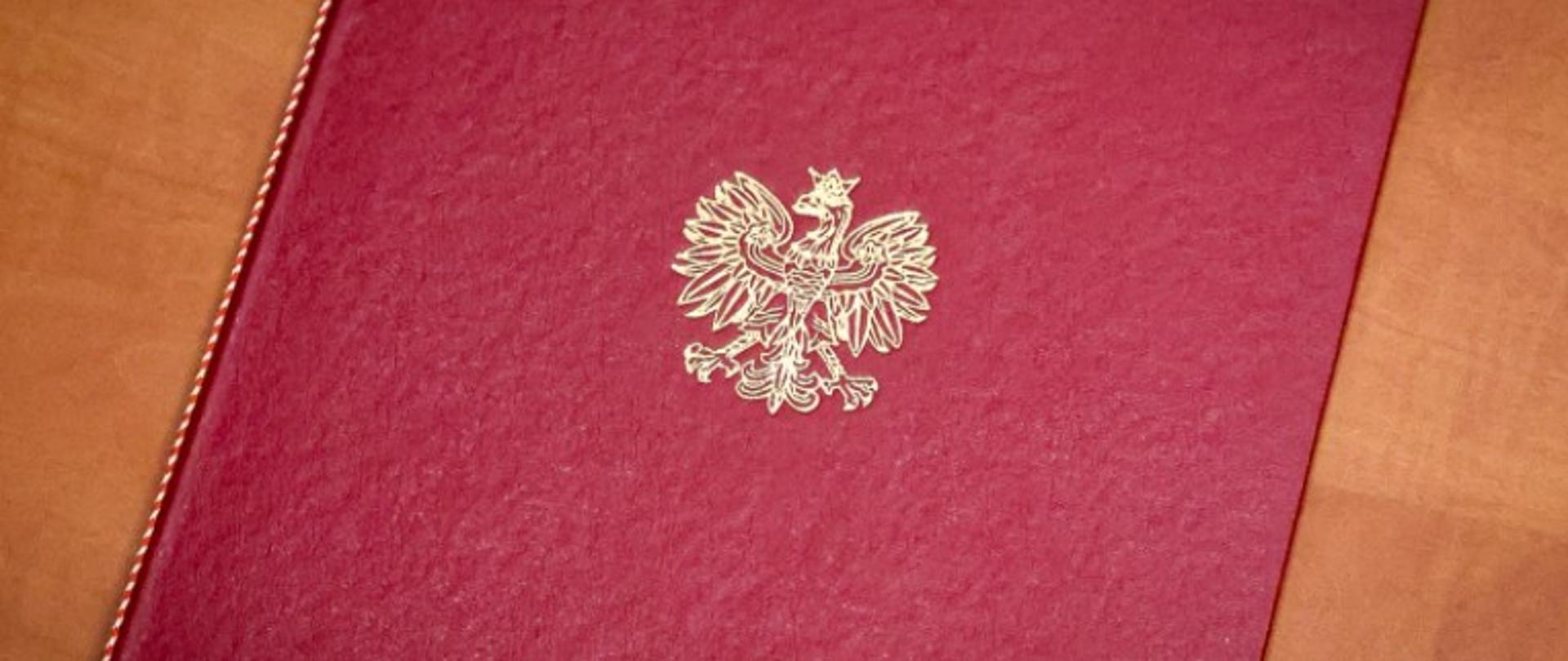 teczka na akta barwy ciemnoczerwonej z biegnącą wzdłuż grzbietu ozdobną nitką barwy biało-czerwonej ze wzorem ułożonym na skos oraz logotypem Orła Białego barwy złotej
