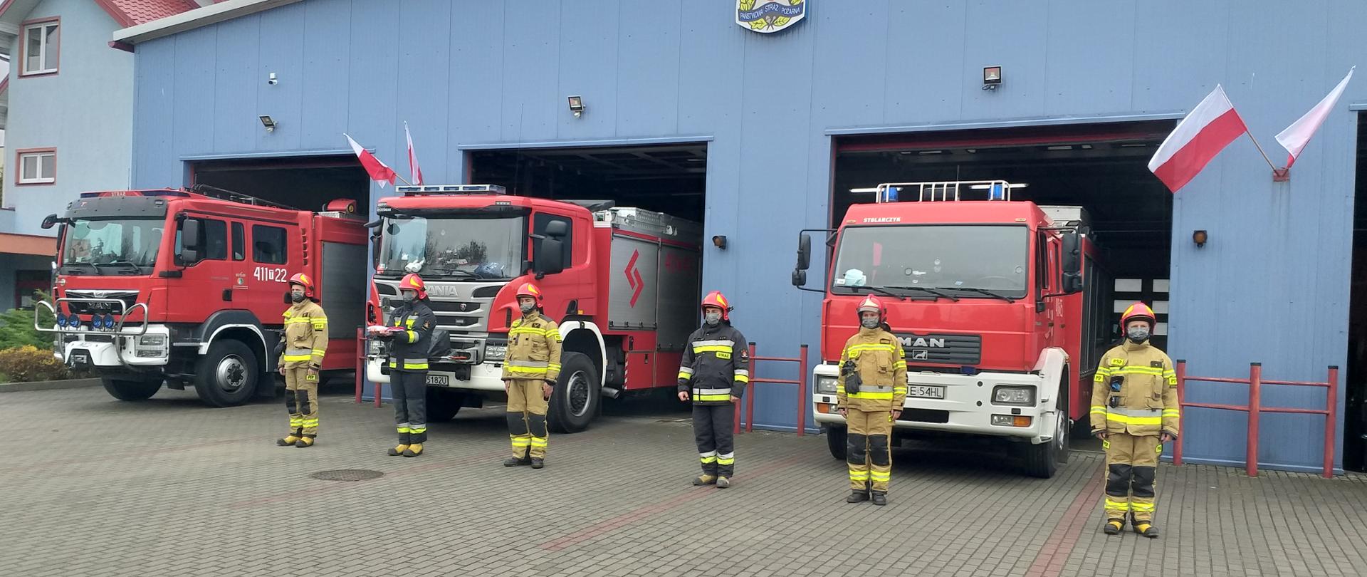 Zdjęcie przedstawia sześciu strażaków w ubraniach specjalnych z założonymi maseczkami ochronnymi na twarz. W tle znajduję się garaż Jednostki Ratowniczo- Gaśniczej z otwartymi bramami. W bramach widoczne trzy samochody ratowniczo- gaśnicze. Drugi strażak od lewej trzyma w rękach flagę biało- czerwoną.