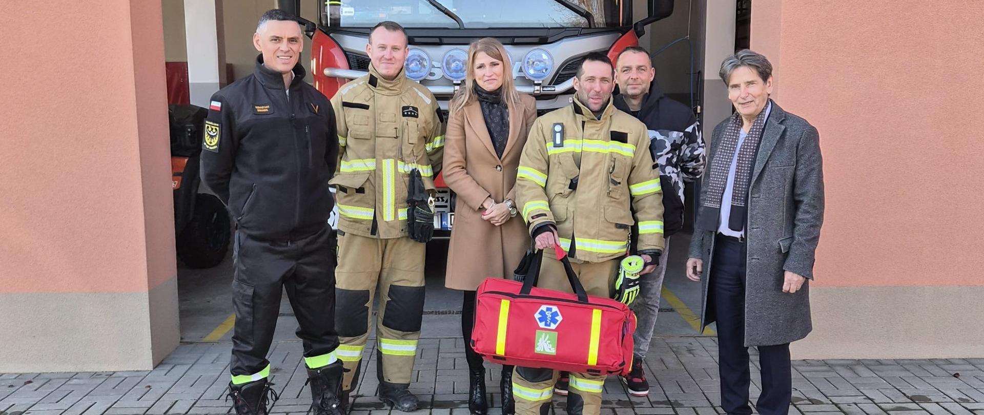 Kobieta i pięciu mężczyzn pozują do zdjęcia na tle garażu, w którym stoi wóz bojowy straży pożarnej. Czterej mężczyźni są w strojach koszarowych OSP. Jeden z nich trzyma w rękach czerwoną torbę z zestawem ratownictwa.