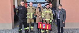 Kobieta i pięciu mężczyzn pozują do zdjęcia na tle garażu, w którym stoi wóz bojowy straży pożarnej. Czterej mężczyźni są w strojach koszarowych OSP. Jeden z nich trzyma w rękach czerwoną torbę z zestawem ratownictwa.