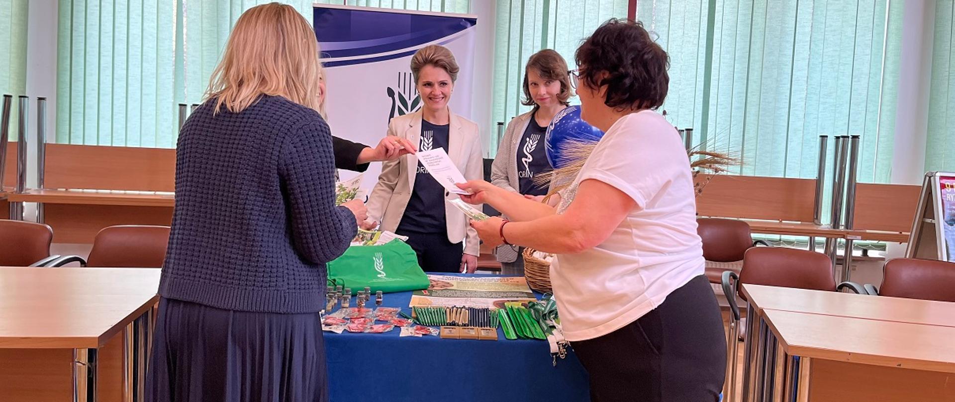 Trzy panie inspektor przedstawiają stoisko promujące Inspekcję. Za nimi baner z logo Inspekcji. Na stoisku przed nimi znajdują się gadżety i materiały edukacyjne: długopisy, ołówki, kredki, opaski odblaskowe, breloczki i worki. Dwie Panie odwiedzające stoisko dostają ulotki informacyjne. 