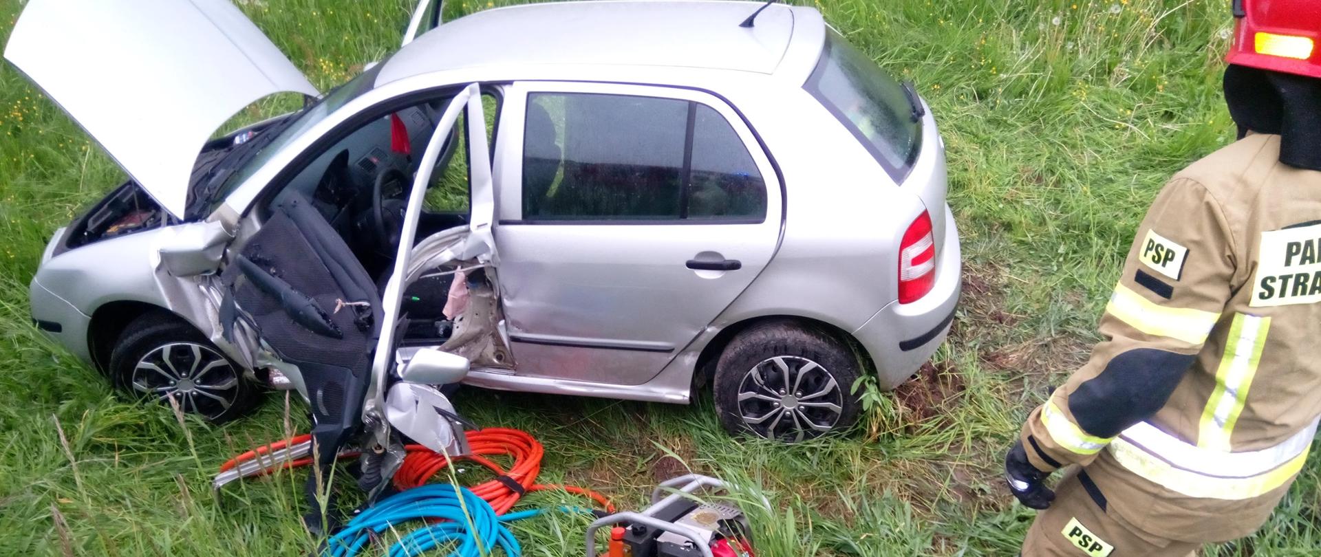 Widoczny strażak na dalszym planie sprzęt hydrauliczny oraz uszkodzony samochód osobowy Skoda Fabia