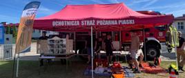 Stanowisko przygotowane przez straż pożarną. Pod namiotem stoi mobilny symulator zagrożeń pożarowych, leżą narzędzia hydrauliczne oraz fantomy. Za namiotem stoi wóz strażacki.
