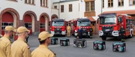 Widok na plac. Tyłem do obiektywu stoją strażacy w szeregu, widać ich trzech. Przed nimi w szeregu 5 agregatów prądotwórczych, dalej 5 samochodów pożarniczych, a na środku mównica i przemawiający wojewoda. W tle budynki strażnicy.