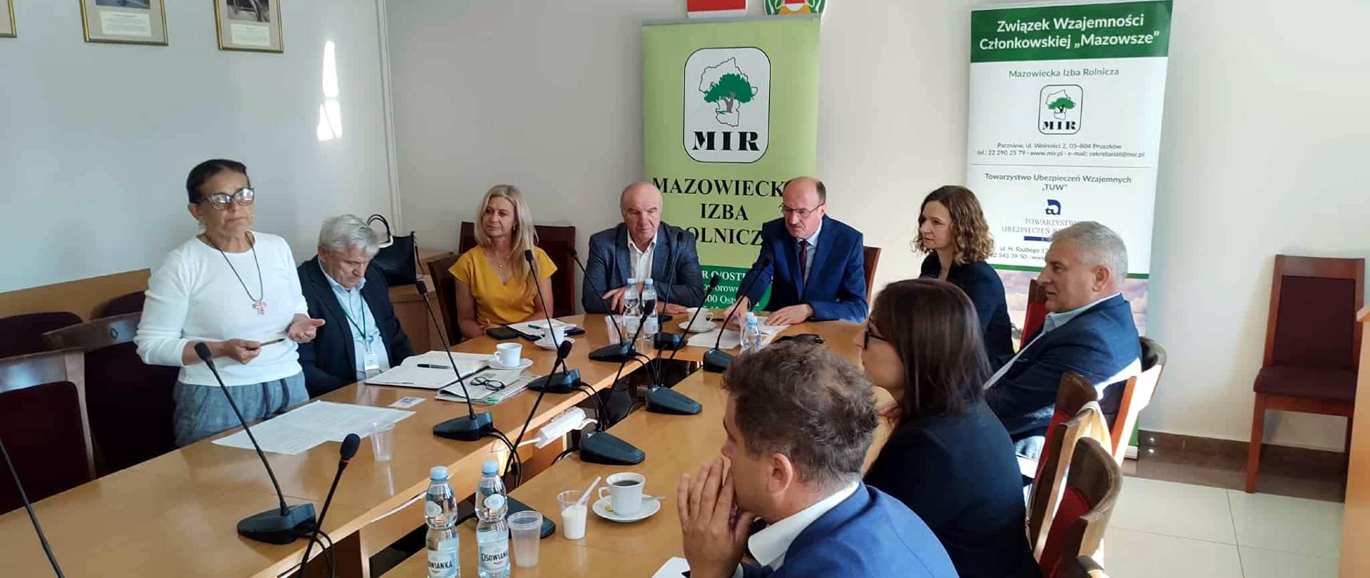 Grupa osób siedzi przy stole konferencyjnym podczas spotkania. Na stole znajdują się mikrofony, dokumenty, filiżanki z kawą i butelki wody. W tle widoczne są banery z logo Mazowieckiej Izby Rolniczej oraz godło Polski i krzyż na ścianie. Jedna z osób, kobieta w białym swetrze, stoi i przemawia do pozostałych uczestników.