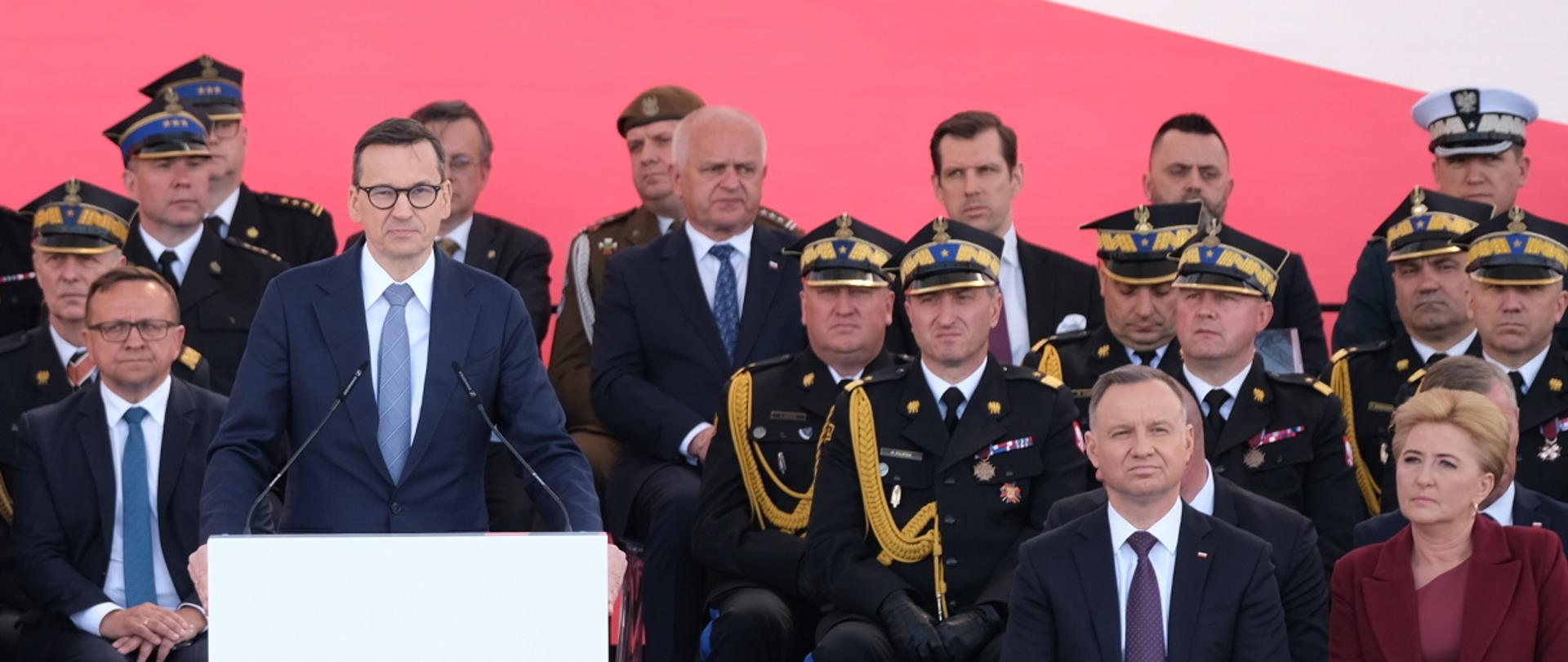 Widok z góry na strażaków stojących na placu oraz na trybunę z gośćmi, przemawia do mikrofonu premier RP