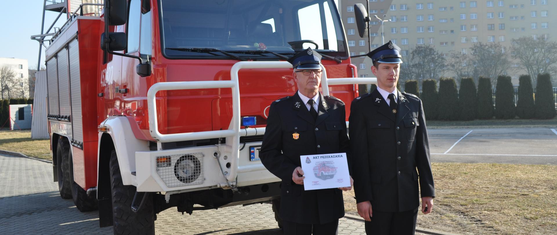 Prezes OSP Bałtów trzyma w rękach akt przekazania auta na tle otrzymanego średniego samochodu gaśniczego. 