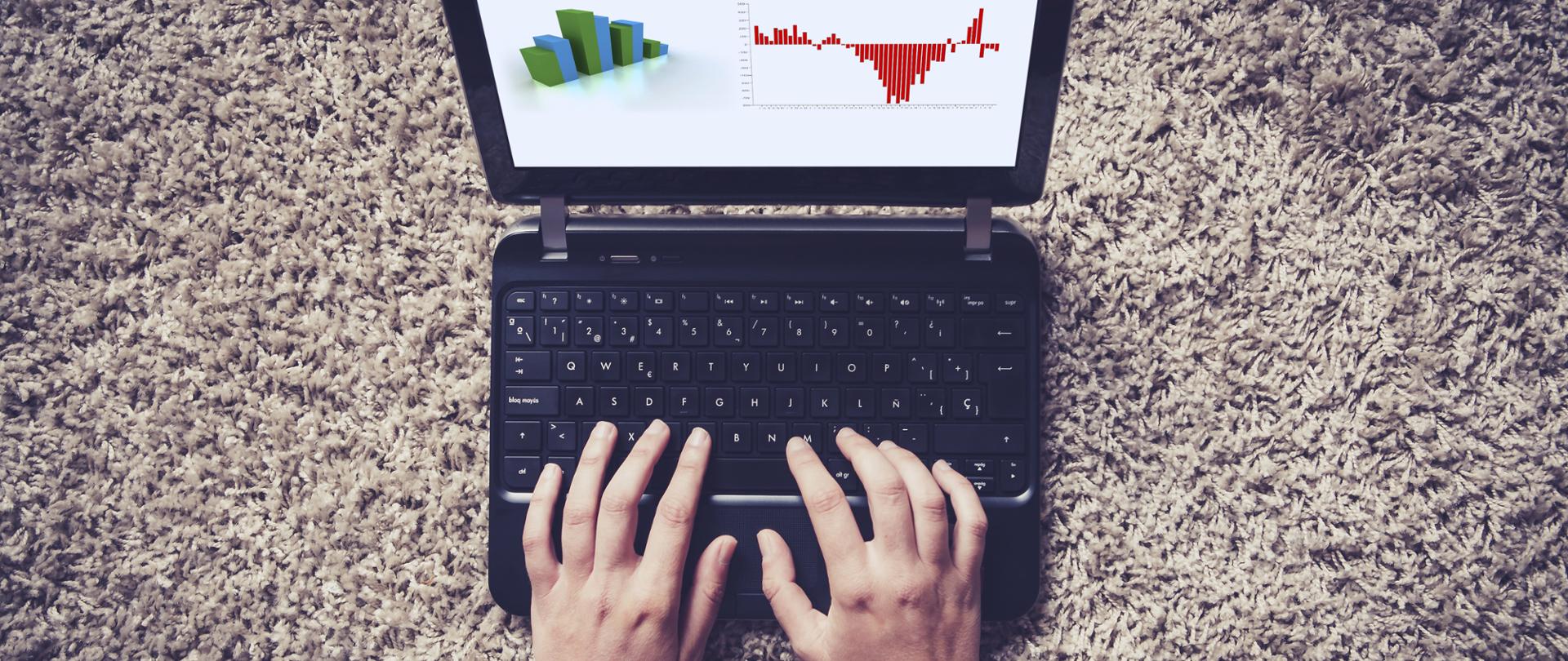 Ręce dotykają klawiatury laptopa leżącego na szarym dywanie