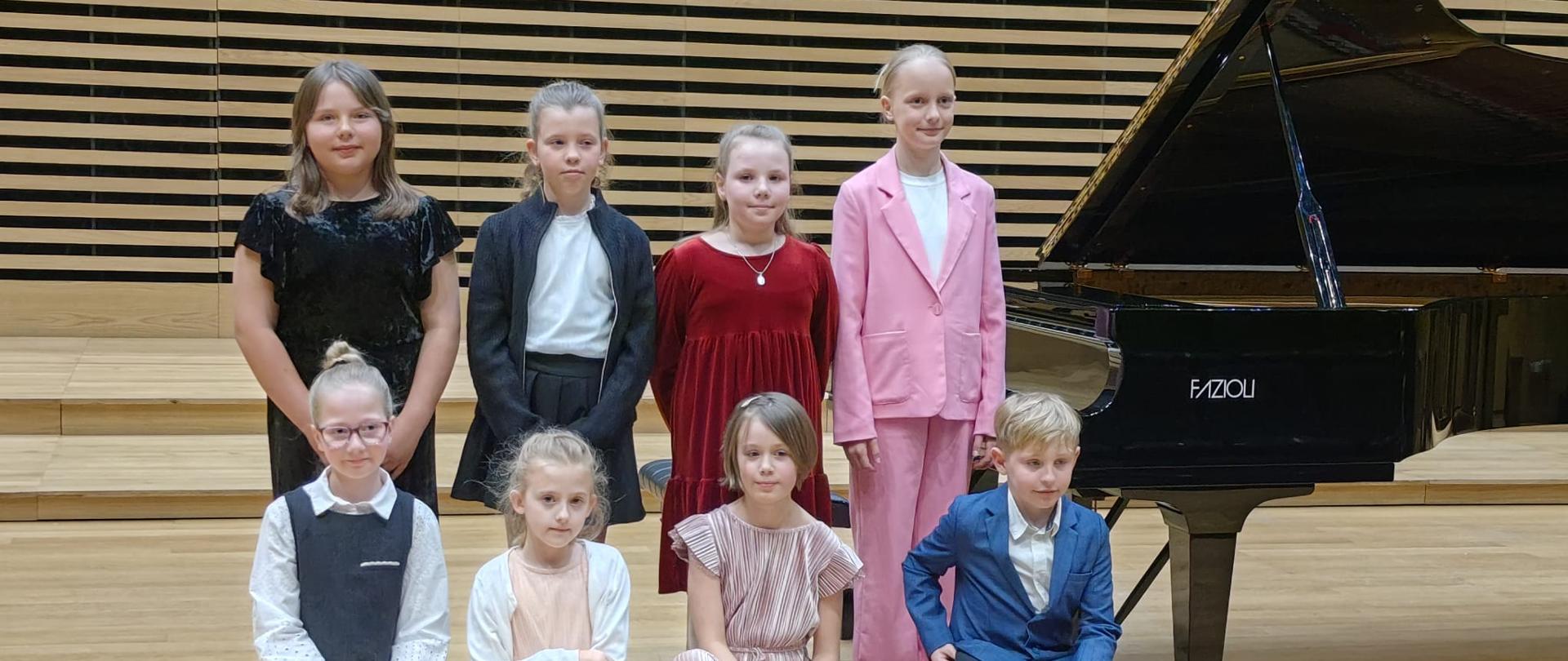 Siedem dziewczynek i chłopiec stoją i kucają przy fortepianie na scenie.