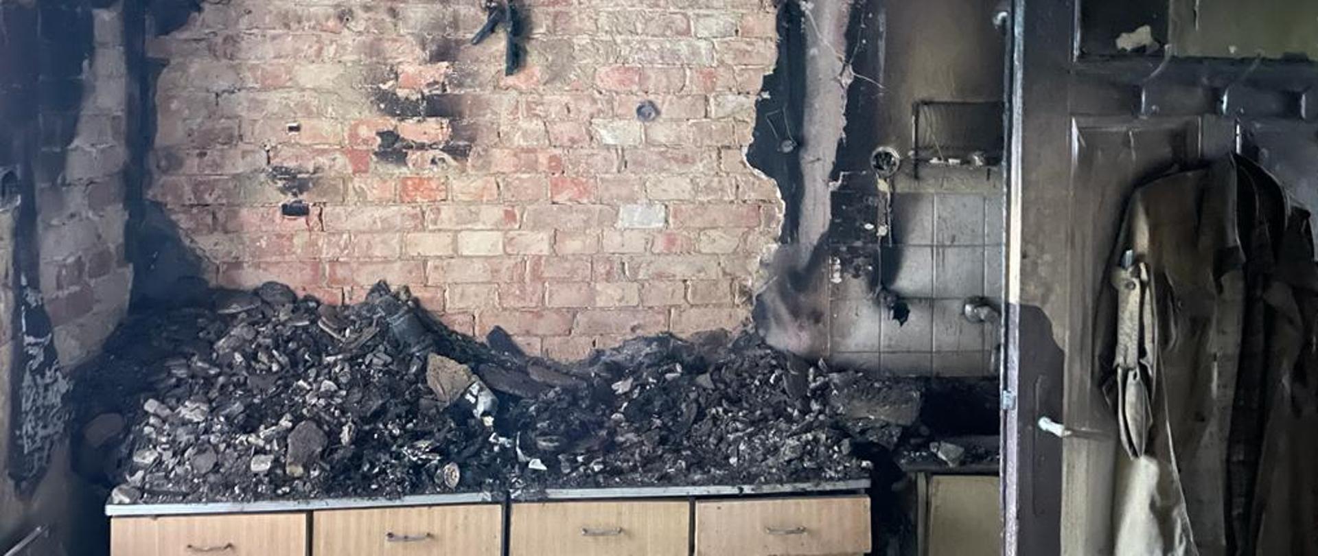 Zdjęcie zrobione wewnątrz spalonego pomieszczenia mieszkania. W centralnej części szafki kuchenne na tle ceglanej ściany, z której odpadł tynk. Po prawej stronie widać otwarte do wewnątrz drzwi na których wiszą nadpalone ubrania.