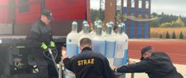 Na zdjęciu widać trzech strażaków, którzy przy pomocy paleciaka próbują załadować skrzynię z butlami z tlenem na samochód kontenerowy. W tle wspinalnia do drabiny hakowej.