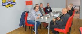 Narada z kominiarzami i zarządcami budynków wielorodzinnych. Świetlica KP PSP w Rawiczu. Przy wspólnym stole siedzi trzech funkcjonariuszy komendy, a na przeciwko nich czterech mężczyzn . Dwaj z nich to mistrzowie kominiarscy, pozostali - zarządcy budynków wielorodzinnych w Rawiczu. W tle na ścianie logo PSP i napis "Komenda Powiatowa PSP w Rawiczu" Na stole filiżanki z kawą, ciastka, talerzyki.