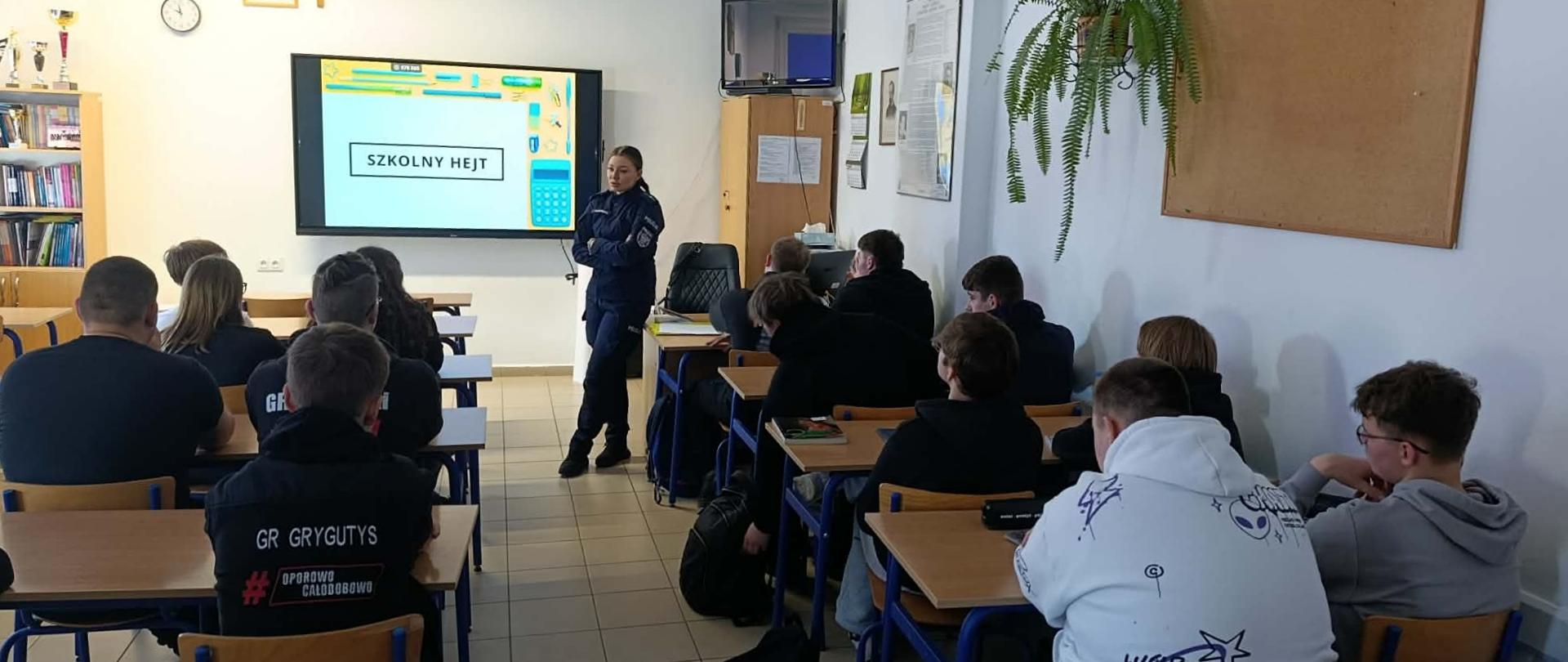 Uczniowie siedzą w ławkach, policjantka prowadzi prelekcję, na monitorze multimedialnym wyświetlona jest prezentacja pt. Szkolny hejt.