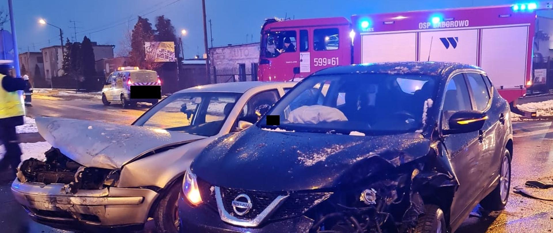Dwa uszkodzone samochody na skrzyżowaniu, w tle widać samochód strażacki