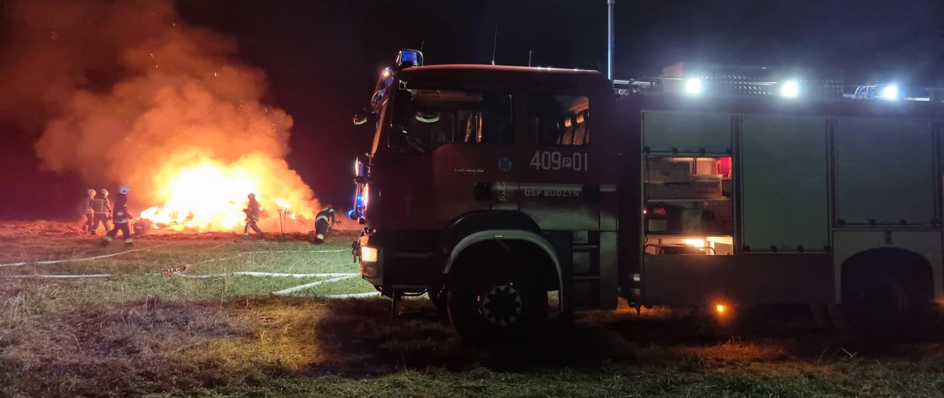 Zdjęcie przedstawia strażaków gaszących pożar stogu słomy.
W tle samochody strażackie oraz pole.
